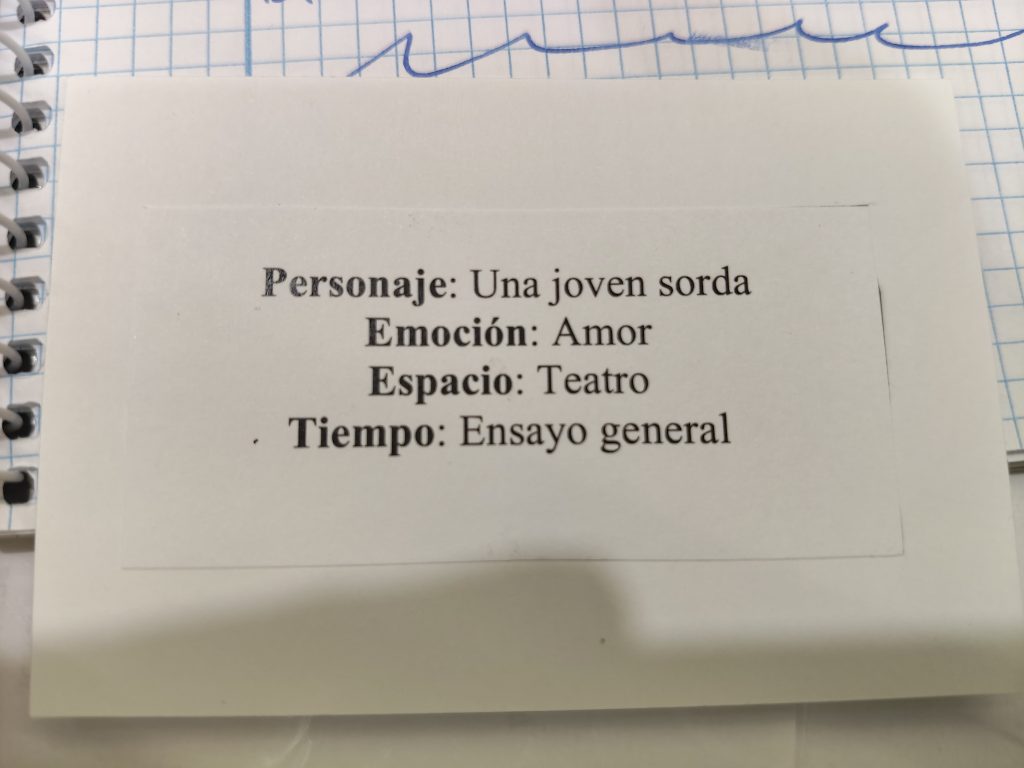 Foto de una tarjeta blanca donde aparecen escrito en negrita, de arriba a abajo, los siguientes textos: 
Personaje: Una joven sorda
Emoción: Amor
Espacio: Teatro
Tiempo: Ensayo general