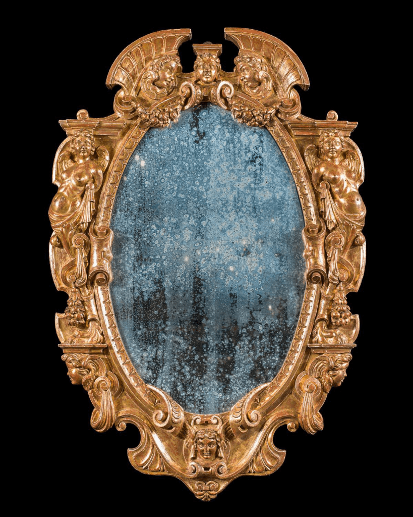 Imagen de un espejo antiguo con los bordes como de bronce. Está decorado con cabezas y cuerpos completos de puttos (ángeles bebés). La superficie reflectante del espejo está muy estropeada por el paso del tiempo.