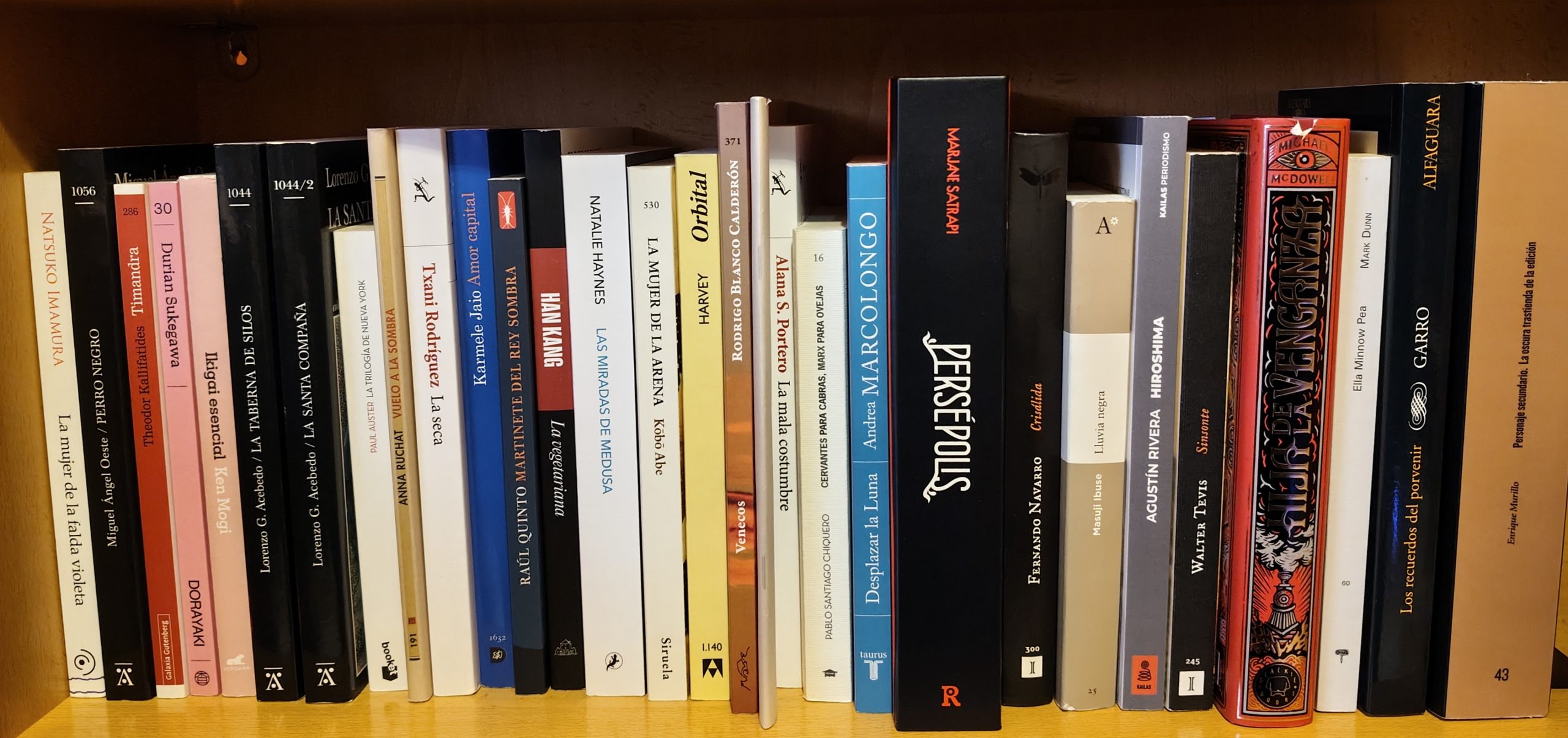 Una serie de libros, y un cómic, puestos en una estantería. Los libros son de diferentes tamaños, colores y formatos, Podemos leer en sus lomos los títulos (de izquierda a derecha): la mujer de la falda violeta, perro negro, timandra, dorayaki, Ikigai esencial, la taberna de silos, la santa compaña, la trilogía de Nueva York, vuelo a la sombra, La seca, amor capital, martinete del rey sombra, la vegetariana, las miradas de Medusa, la mujer de la arena, orbital, venecos, la mala costumbre, Cervantes para cabras Marx para ovejas, desplazar la luna, Persépolis, Crisálida, Lluvia negra, Hirsoshima, Sisonte, Hija de la venganza, Ella Minnow Pea, Los recuerdos del Porvenir y Personaje Secundario.