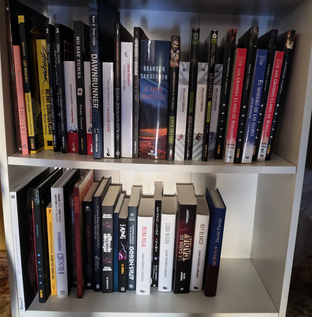 Foto de dos baldas de la hermana más estrecha de la estantería Billie de Ikea. En ella se ven los libros, algún manual y tomos de cómics que tengo pendientes de leer correctamente colocados a lo largo de las dos baldas (vamos, que no están apilados). Se ven alrededor de 45. Los hay muy gordos (como el camino de los reyes o muy finitos como el manual de Warmonger o Carcoma)